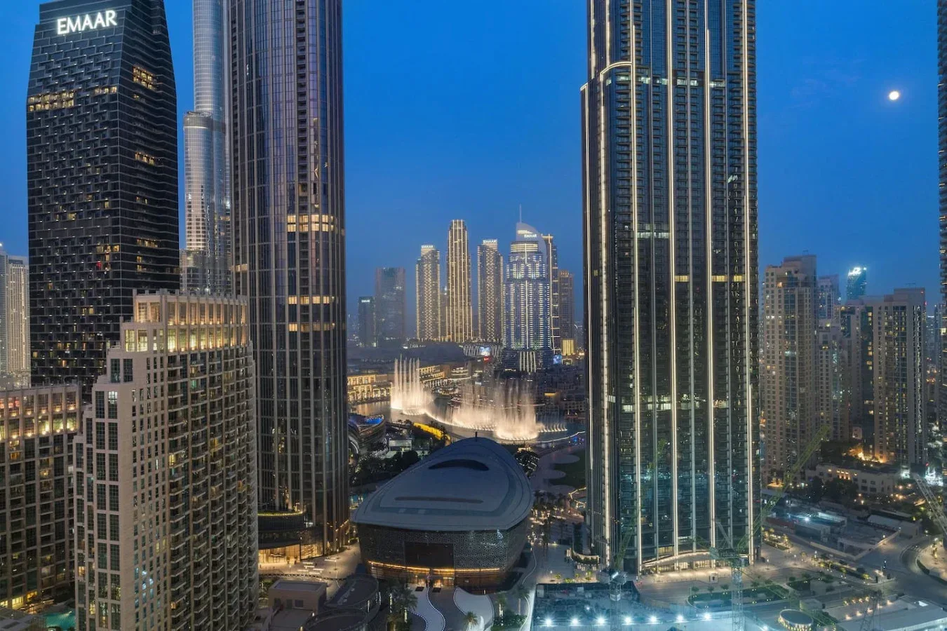A city skyline at dusk, with illuminated skyscrapers and a fountain.