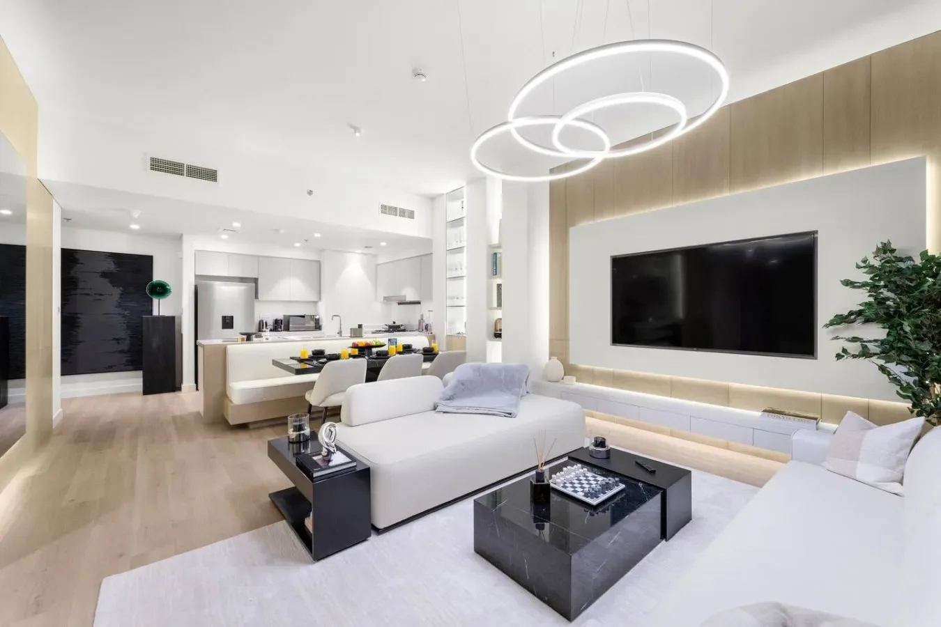 An open-plan living room with white seating, a wall-mounted television, and a kitchen beyond.