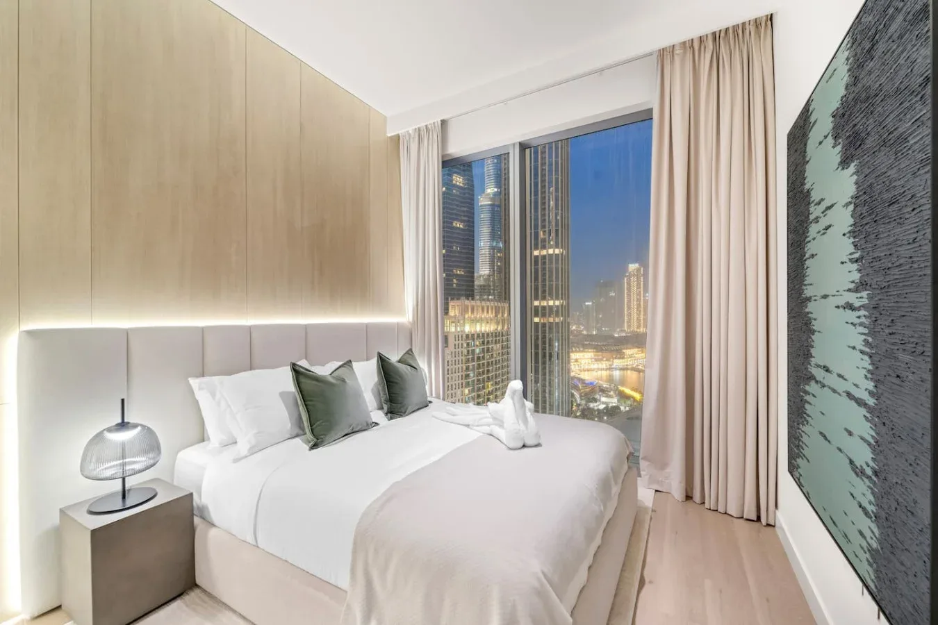 A bedroom with a neatly made bed beside a large window overlooking city lights.