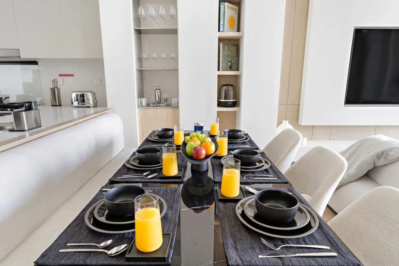 A dining table set with plates and glasses sits beside a bright kitchen and living area.