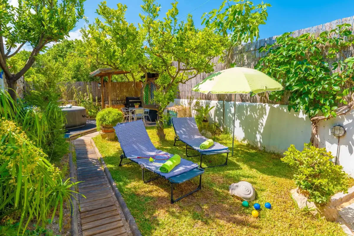 Un patio ajardinado tiene asientos reclinables, una sombrilla y árboles frondosos.
