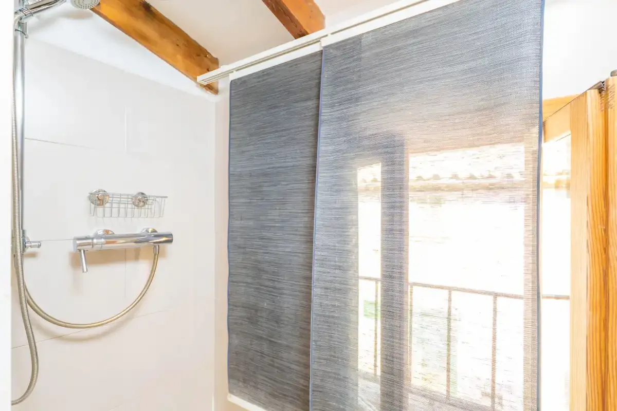 Zona de ducha de un baño con una pantalla gris y una ventana luminosa.
