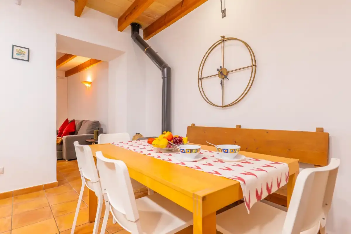 Un comedor con una mesa de madera, sillas blancas y un mantel estampado.