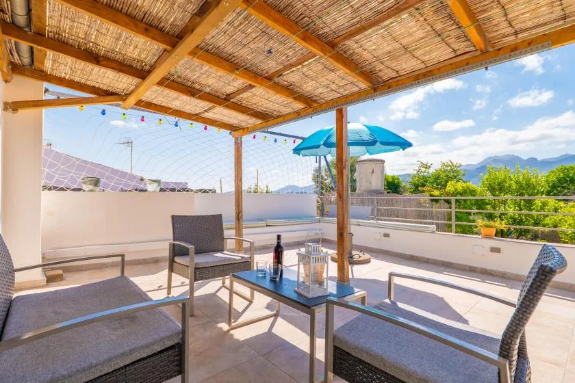 Una terraza exterior con sombra, asientos y una sombrilla azul frente a la vegetación.