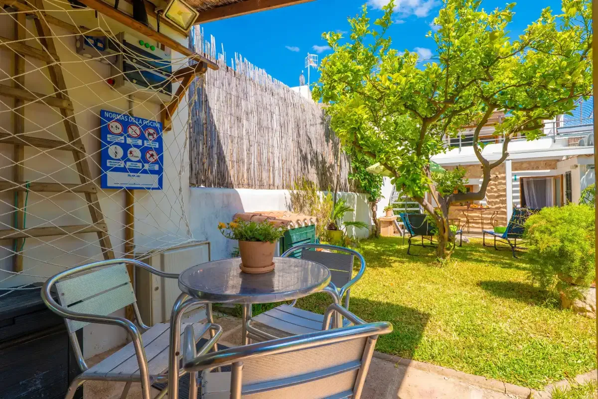 Un patio soleado con asientos metálicos, una mesa y árboles frondosos.