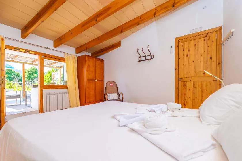 Un dormitorio con una cama blanca, puertas de madera y ventanas con vista al verde.