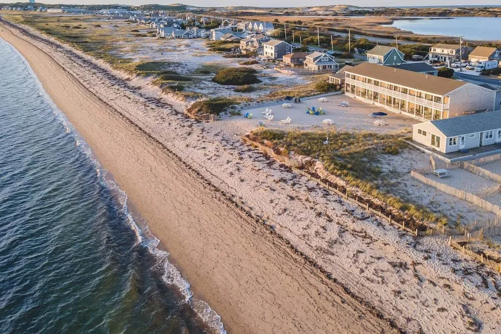 Vista aérea de una playa de arena junto a edificios costeros y aguas poco profundas.