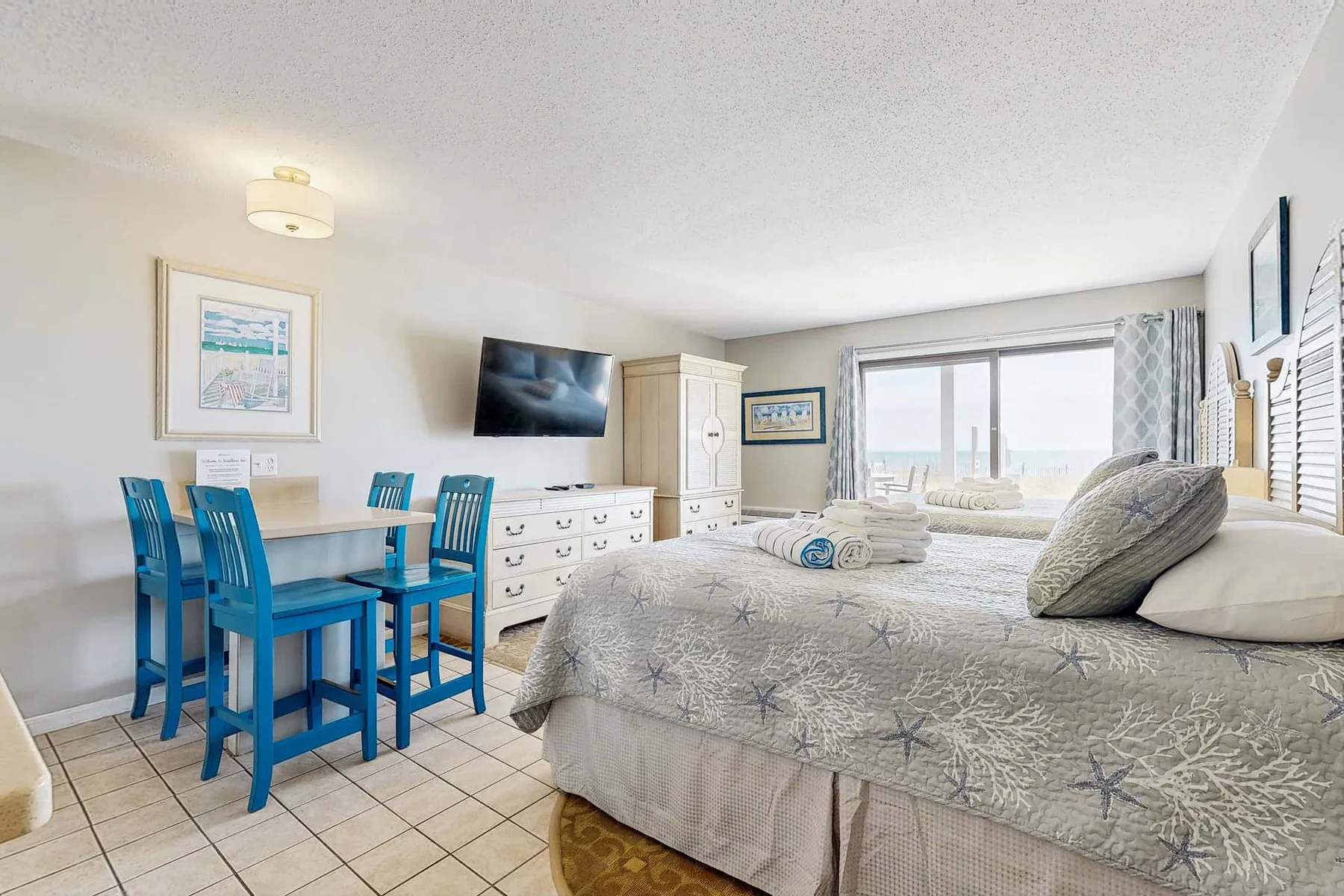Un dormitorio luminoso con una cama, asientos azules y una mesa junto a la ventana.