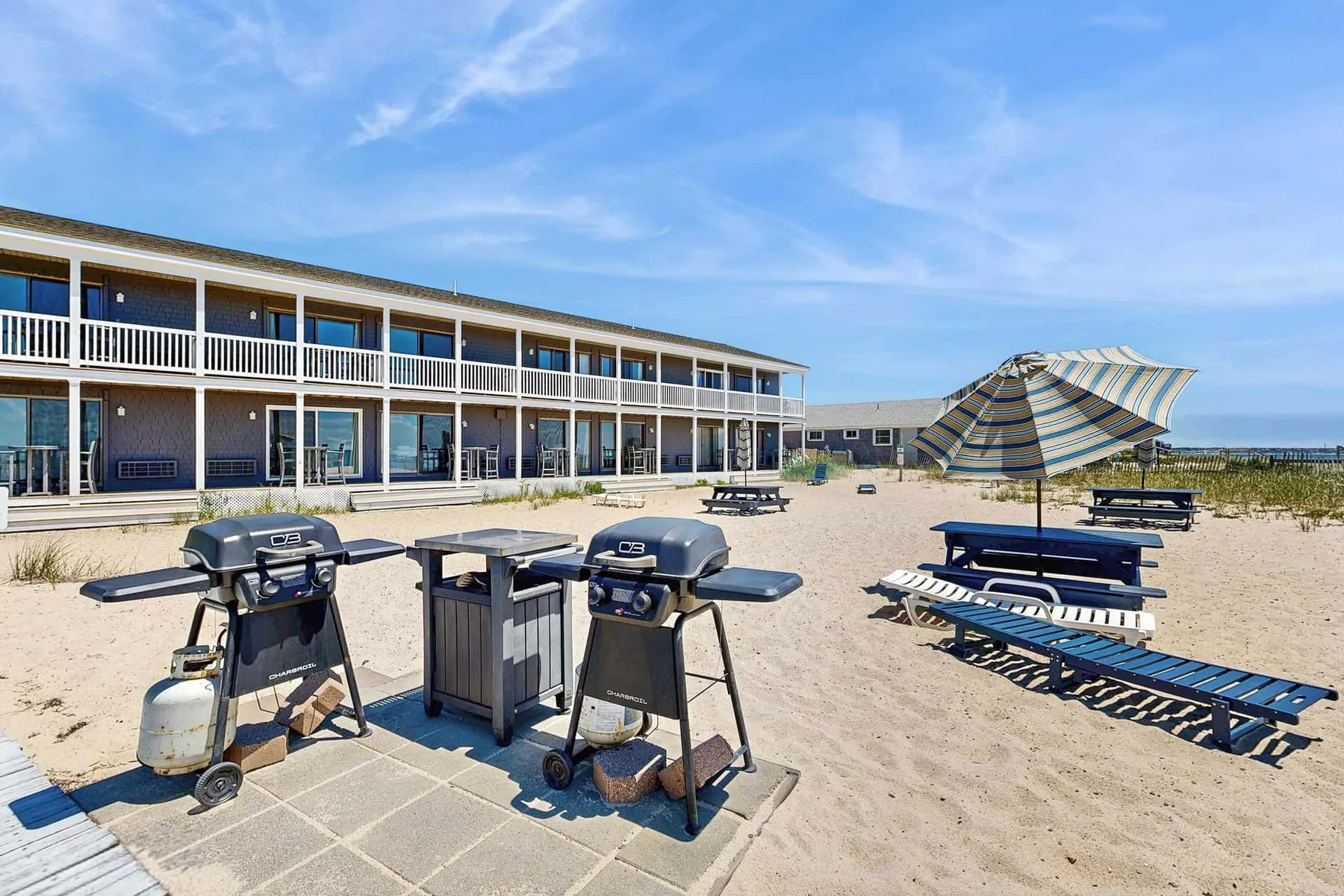 Un patio junto a la playa con parrillas y mesas de picnic junto a un edificio de dos plantas.