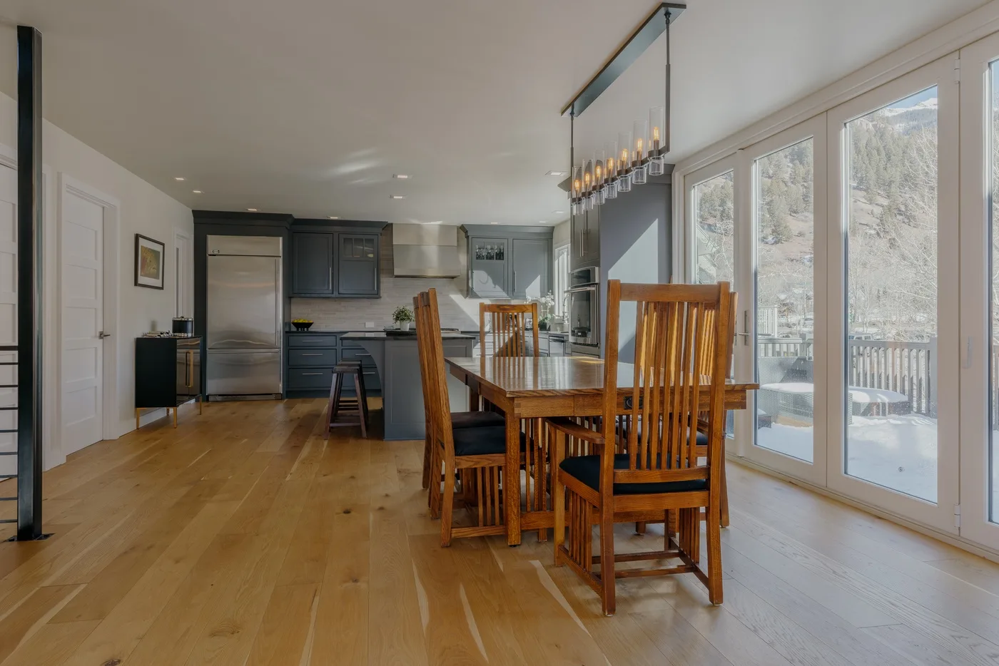 Una mesa de comedor con asientos de madera está junto a una cocina luminosa y grandes ventanas.