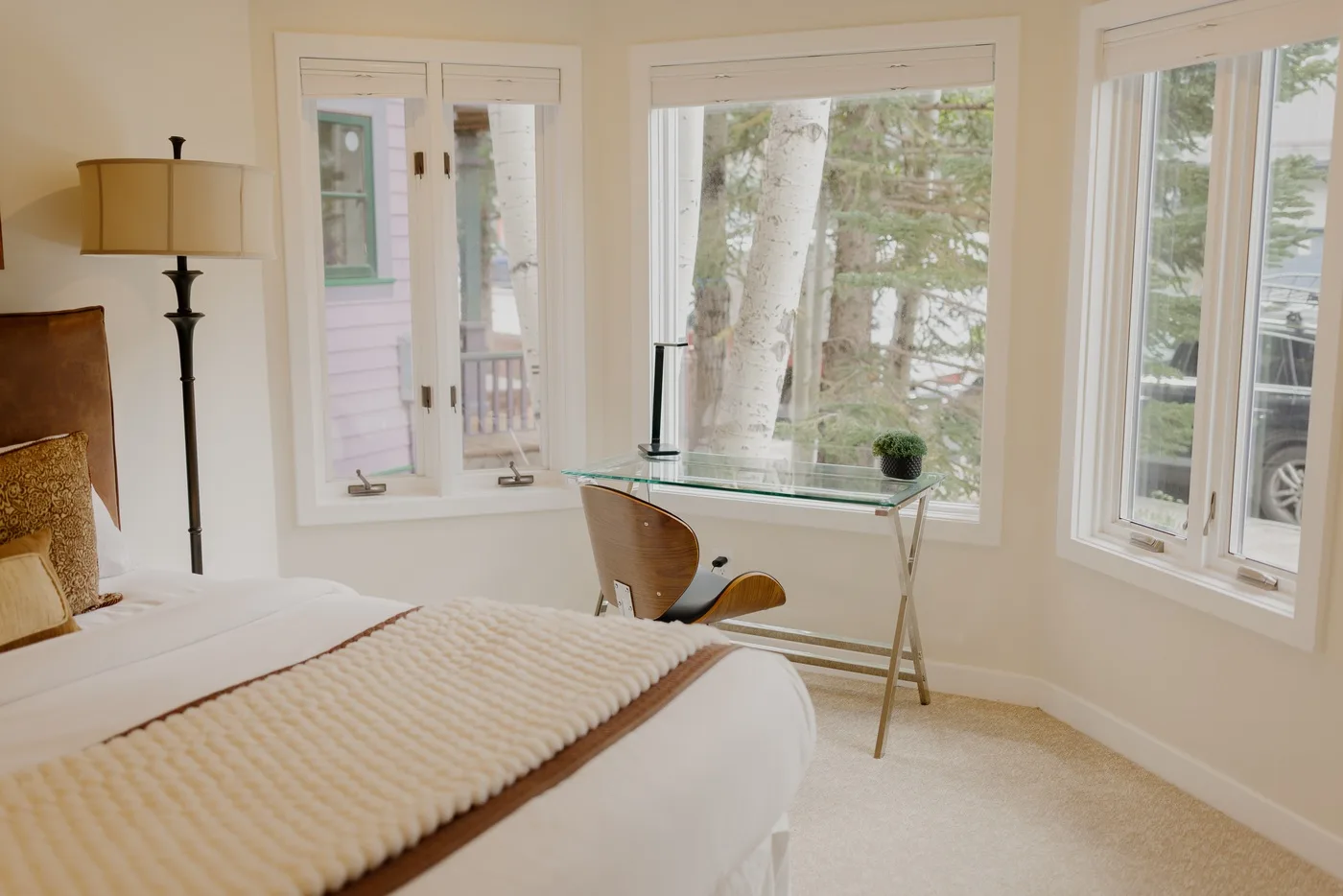 Un dormitorio luminoso con una cama junto a un escritorio con tablero de vidrio y ventanas grandes.