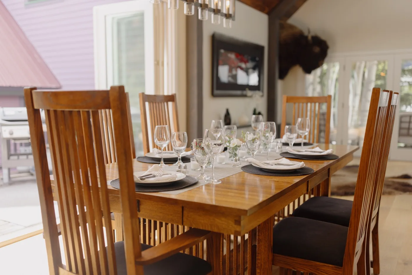Una mesa de comedor de madera está puesta con platos y cristalería en un comedor luminoso.