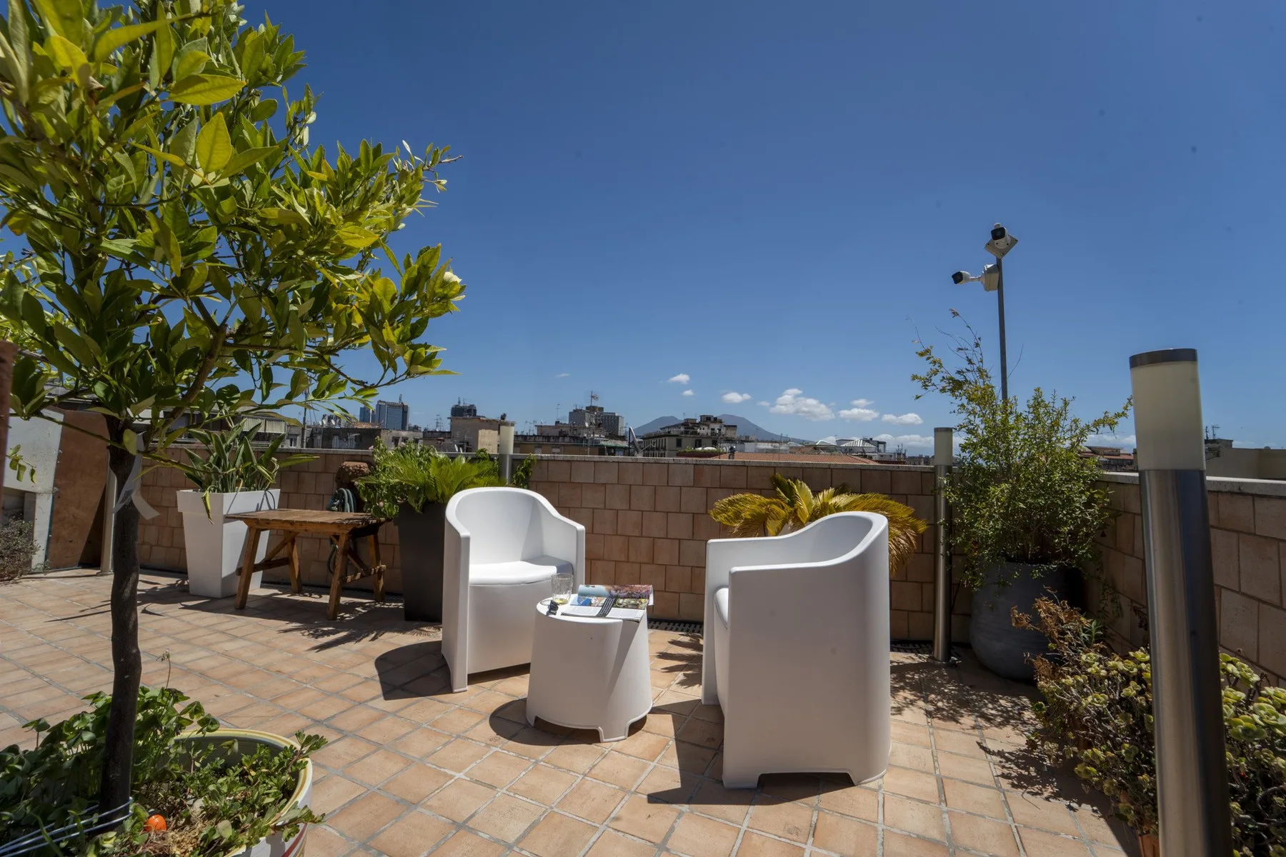 A sunny outdoor terrace has seating, potted plants, and views of nearby buildings.