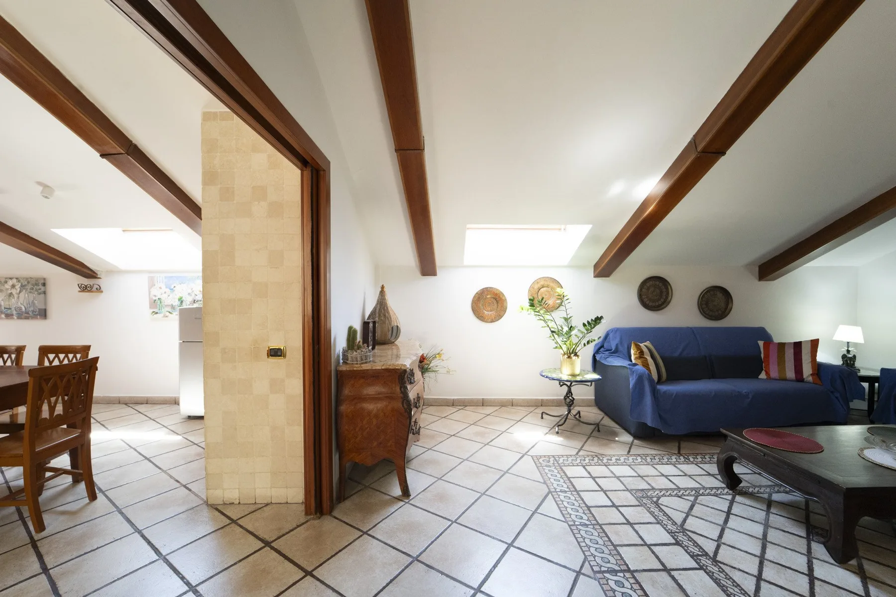 A tiled living room with blue seating, exposed ceiling beams, and a small dining area.