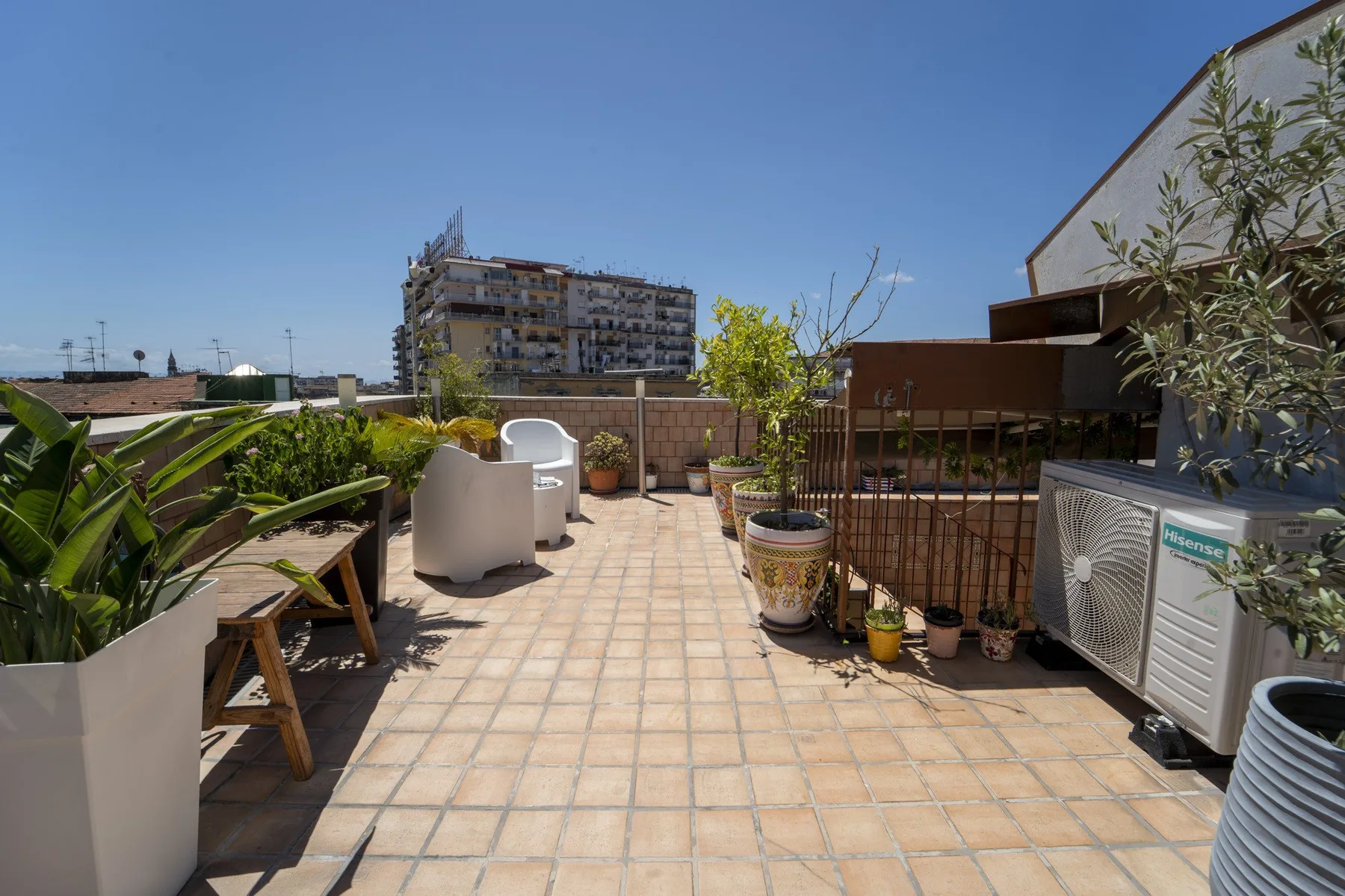 A sunny rooftop terrace with tiled flooring, outdoor seating, and potted plants.