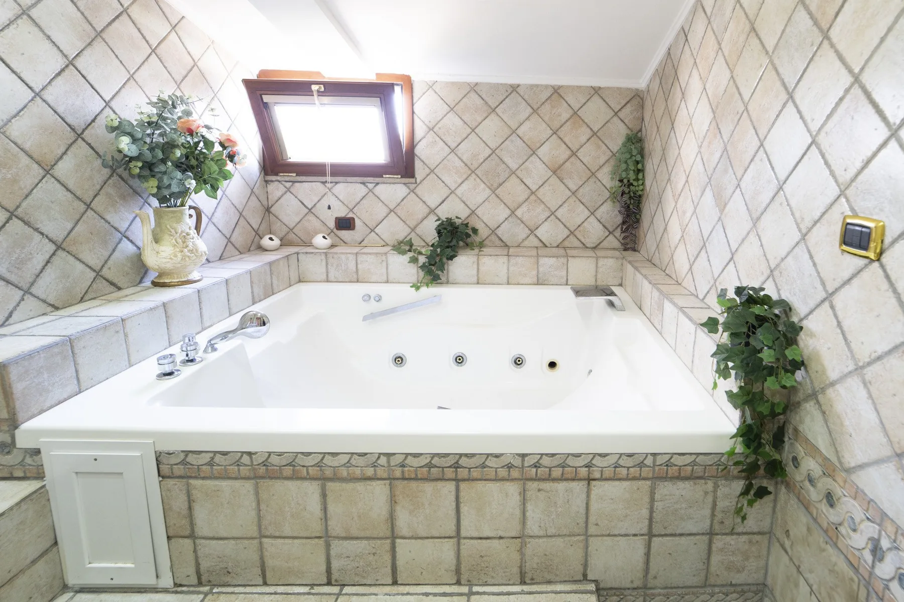 A large bathtub sits beneath a small window in a tiled bathroom.