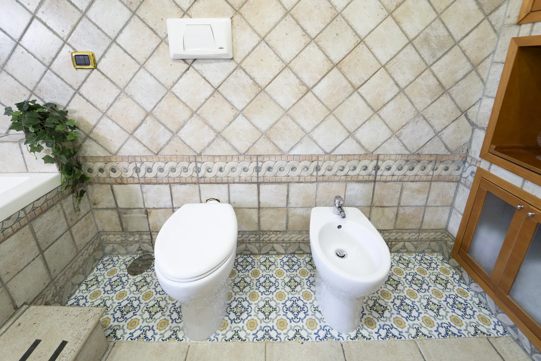 A tiled bathroom with a toilet and bidet beside a wooden cabinet.