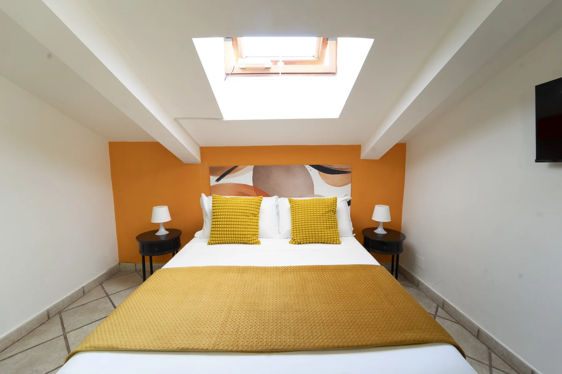 A bedroom with a bed beneath a skylight and orange accent wall.