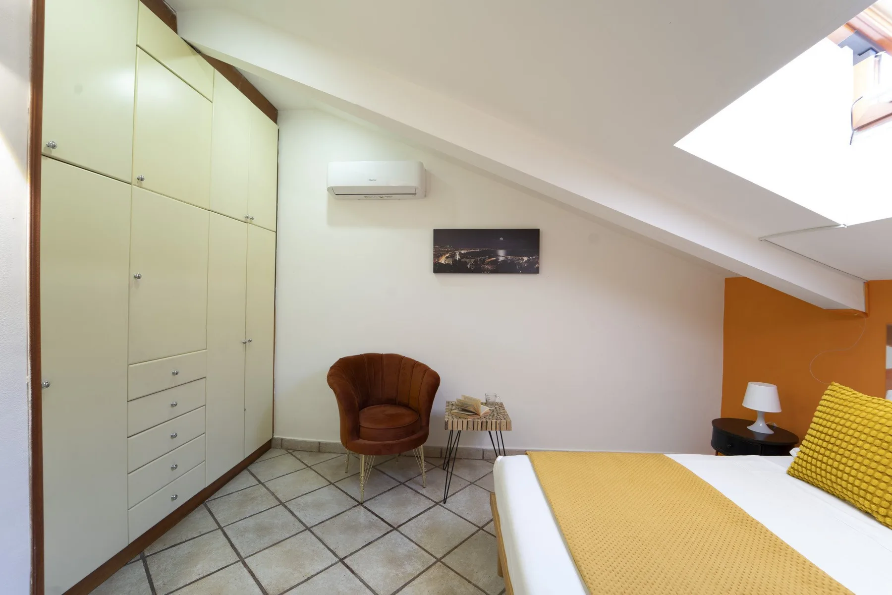 A bedroom with a bed, armchair, and built-in storage beneath a sloped ceiling.