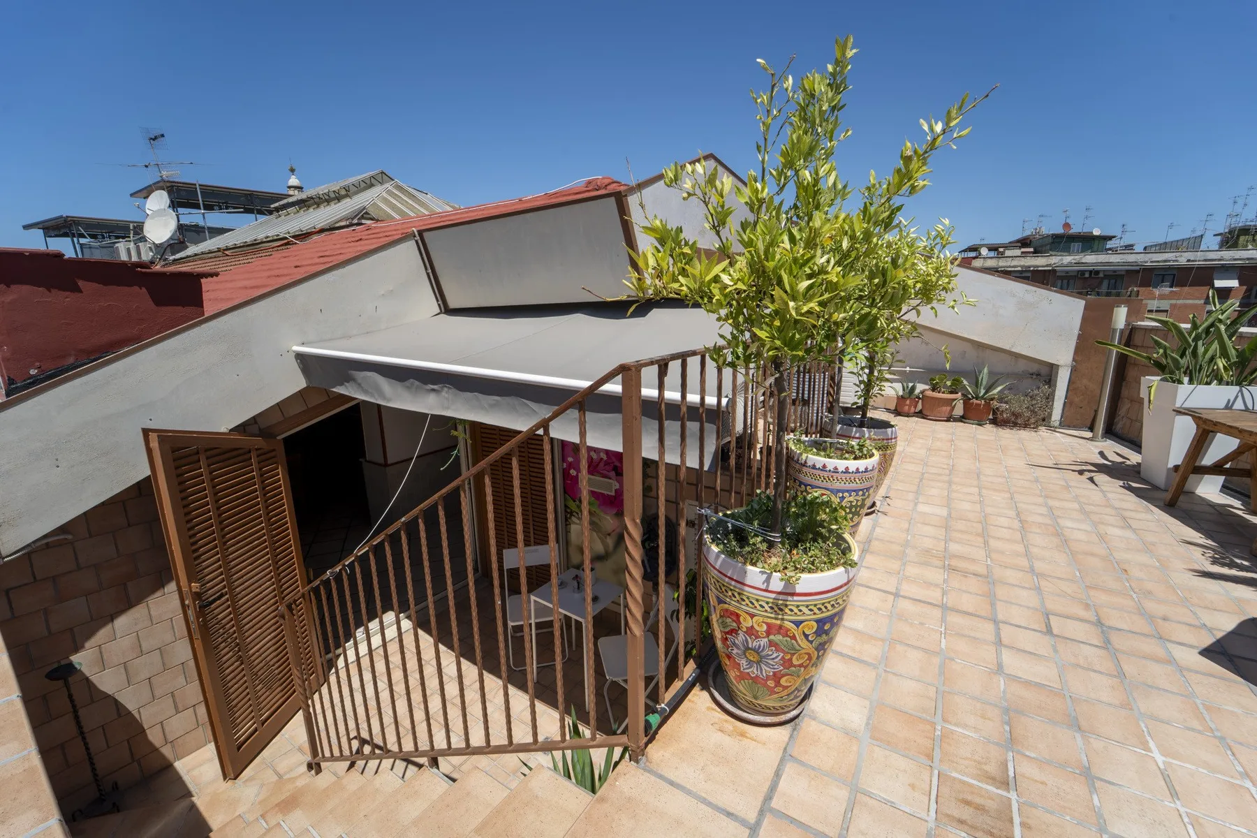 A sunny rooftop terrace with tiled flooring, potted plants, and a shaded seating area.