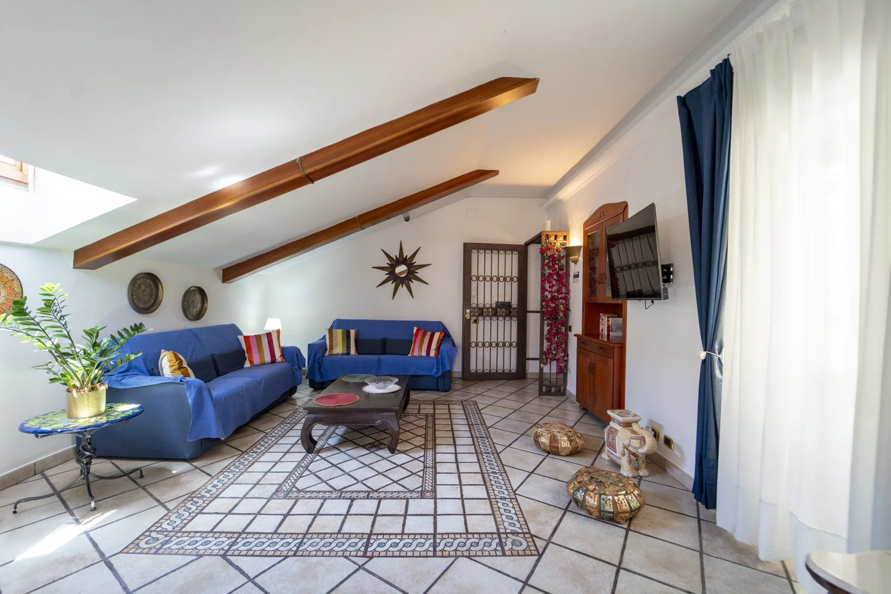 A bright living room with blue seating, a patterned rug, and exposed ceiling beams.