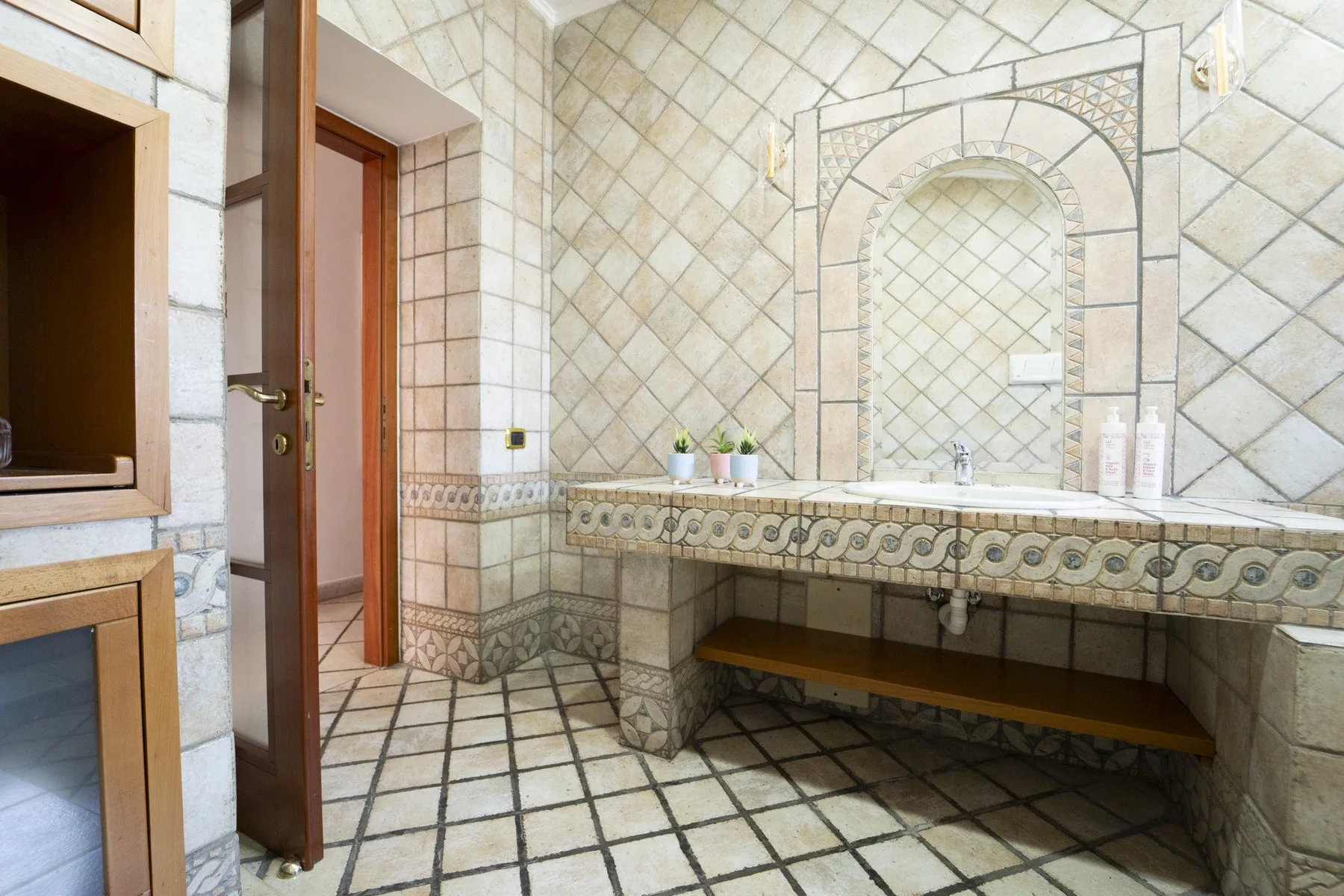 A tiled bathroom with a wide vanity, arched mirror, and open doorway.
