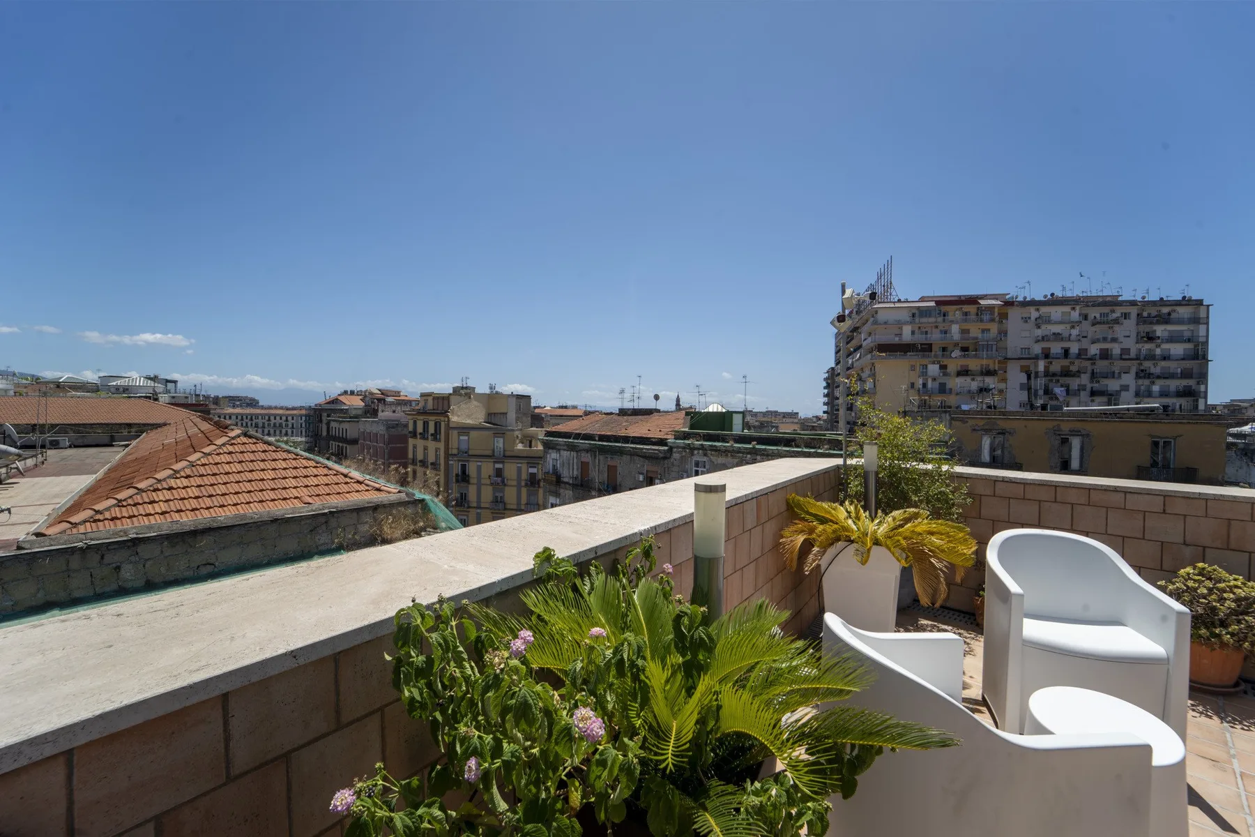 A rooftop terrace overlooks nearby buildings, with white seating and potted plants.