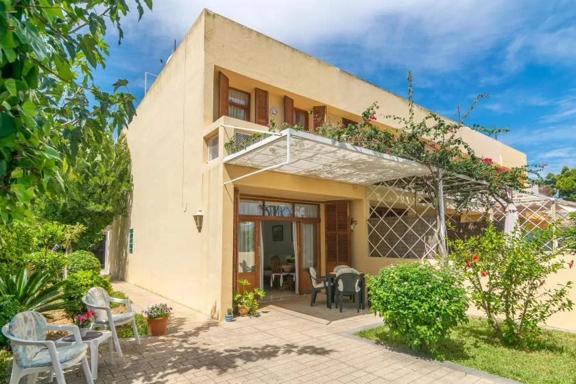 A sunlit house exterior with a leafy garden and a shaded patio.