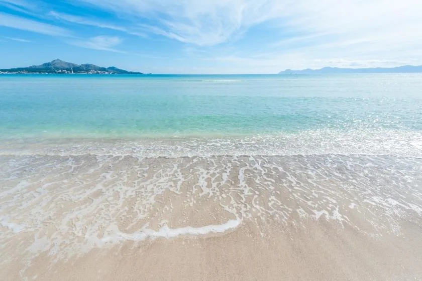 A sandy beach meets clear turquoise water beneath a bright blue sky.