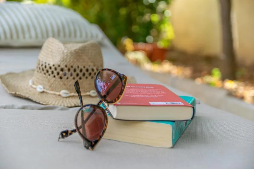 Books and sunglasses rest on an outdoor lounge chair beside a straw hat.