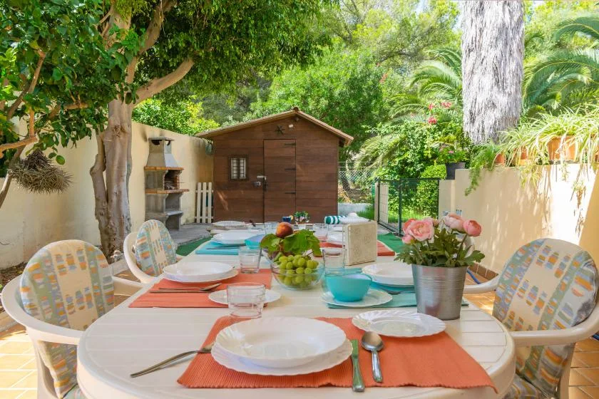 A garden patio dining area with a table set beneath leafy trees.