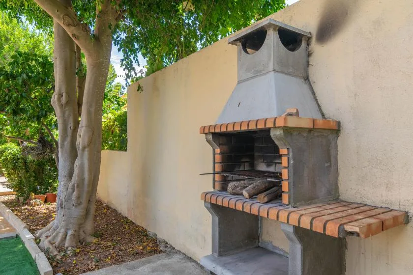 An outdoor brick barbecue stands beside a tree and a pale wall.