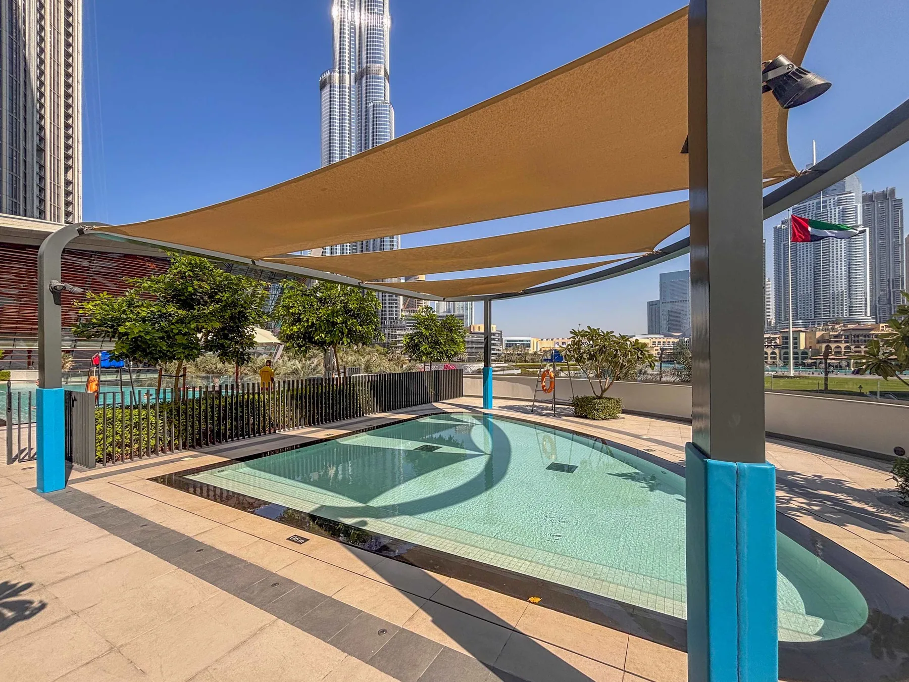Una piscina al aire libre está bajo toldos de sombra, con edificios altos al fondo.
