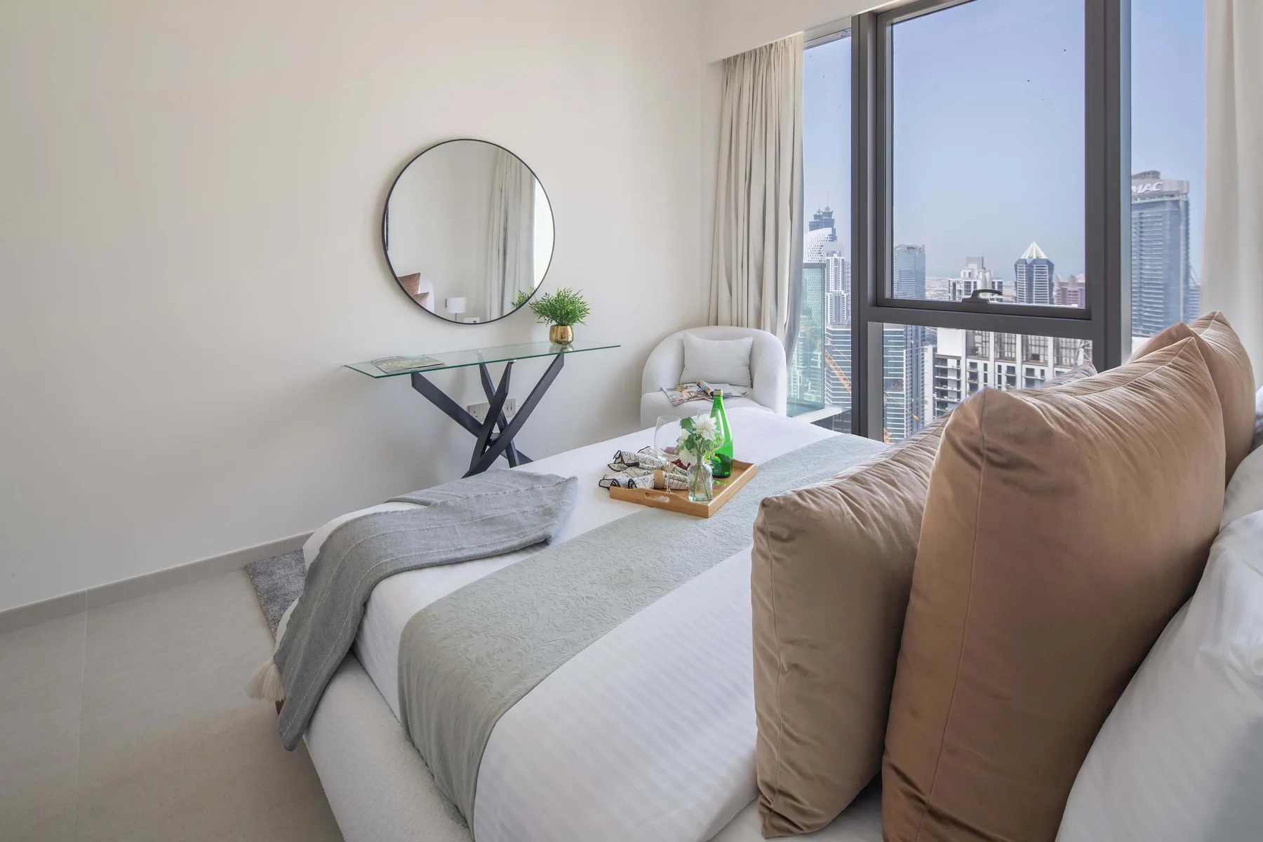 Un dormitorio luminoso con cama, mesa auxiliar y grandes ventanas con vistas a edificios de la ciudad.