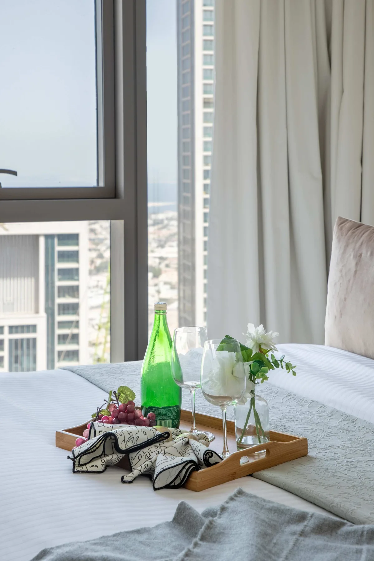 Un dormitorio luminoso con una bandeja de bebidas y flores sobre la cama junto a la ventana.