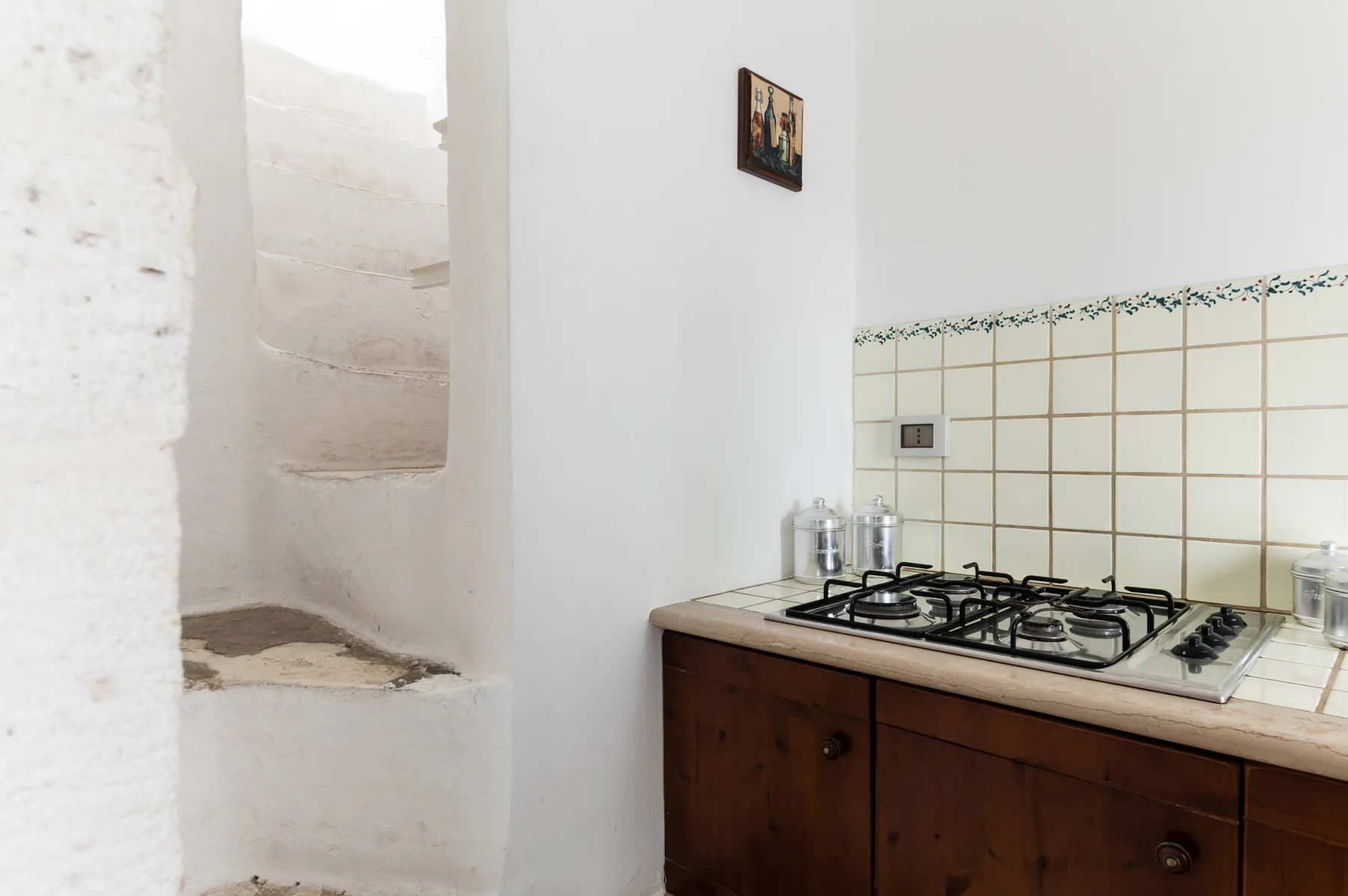 A kitchen with a tiled counter and gas cooktop beside a curved white stairway.