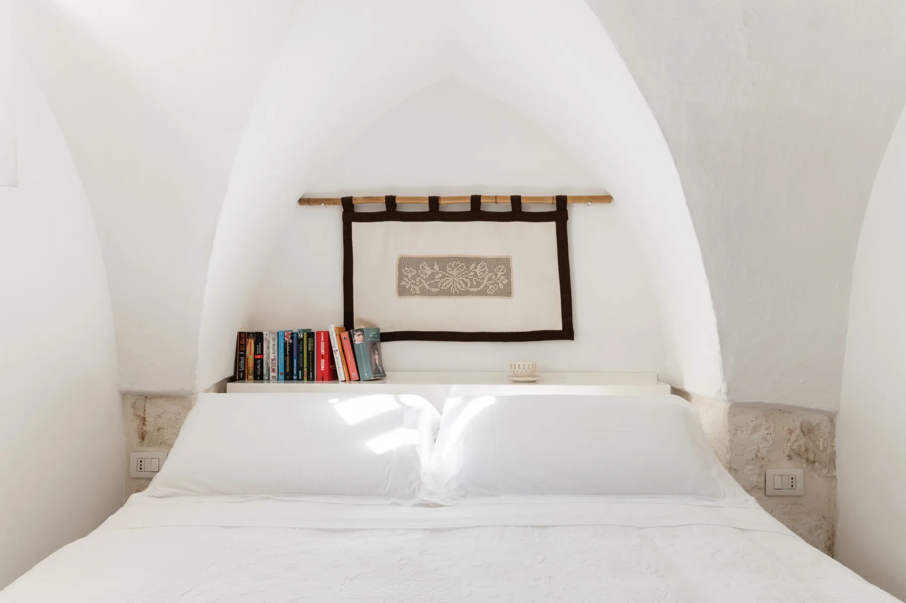A white bed sits beneath an arched ceiling, with books beside it.