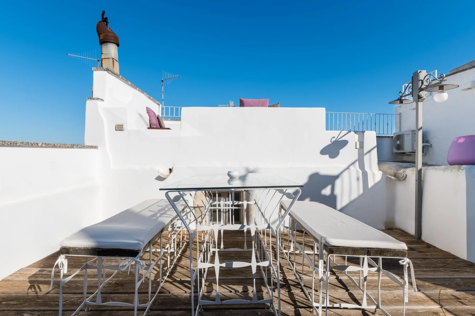 A sunlit rooftop terrace has a dining table and benches against white walls.