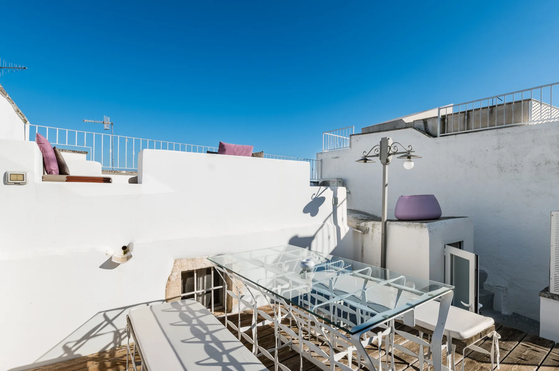 A sunny rooftop terrace with white walls, outdoor seating, and a glass canopy.