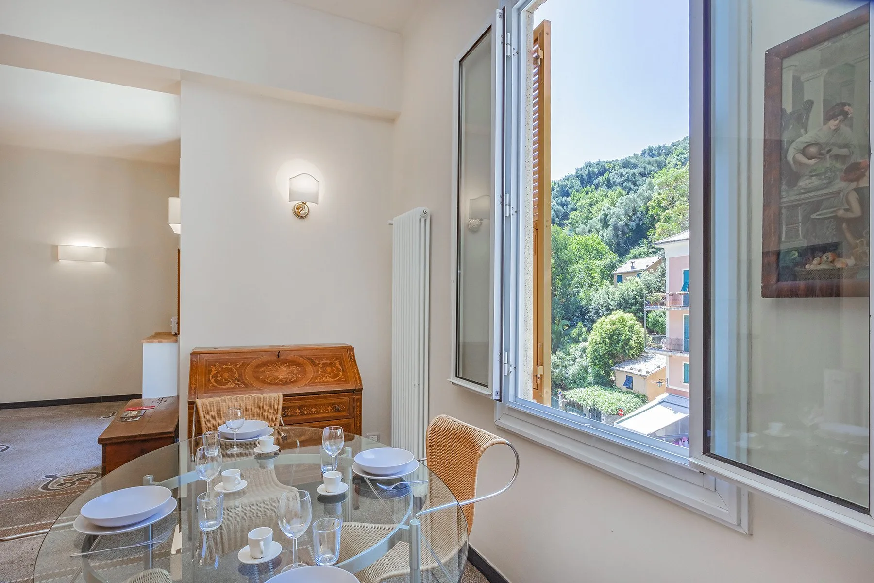A bright dining area with a glass table beside an open window overlooking green hills.