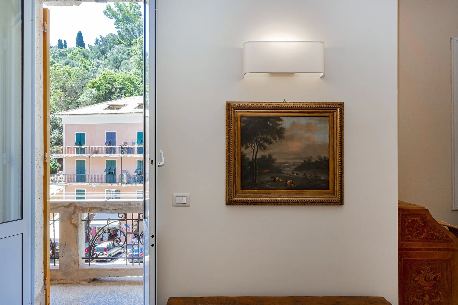 A framed landscape painting hangs beneath a wall light in a bright interior.