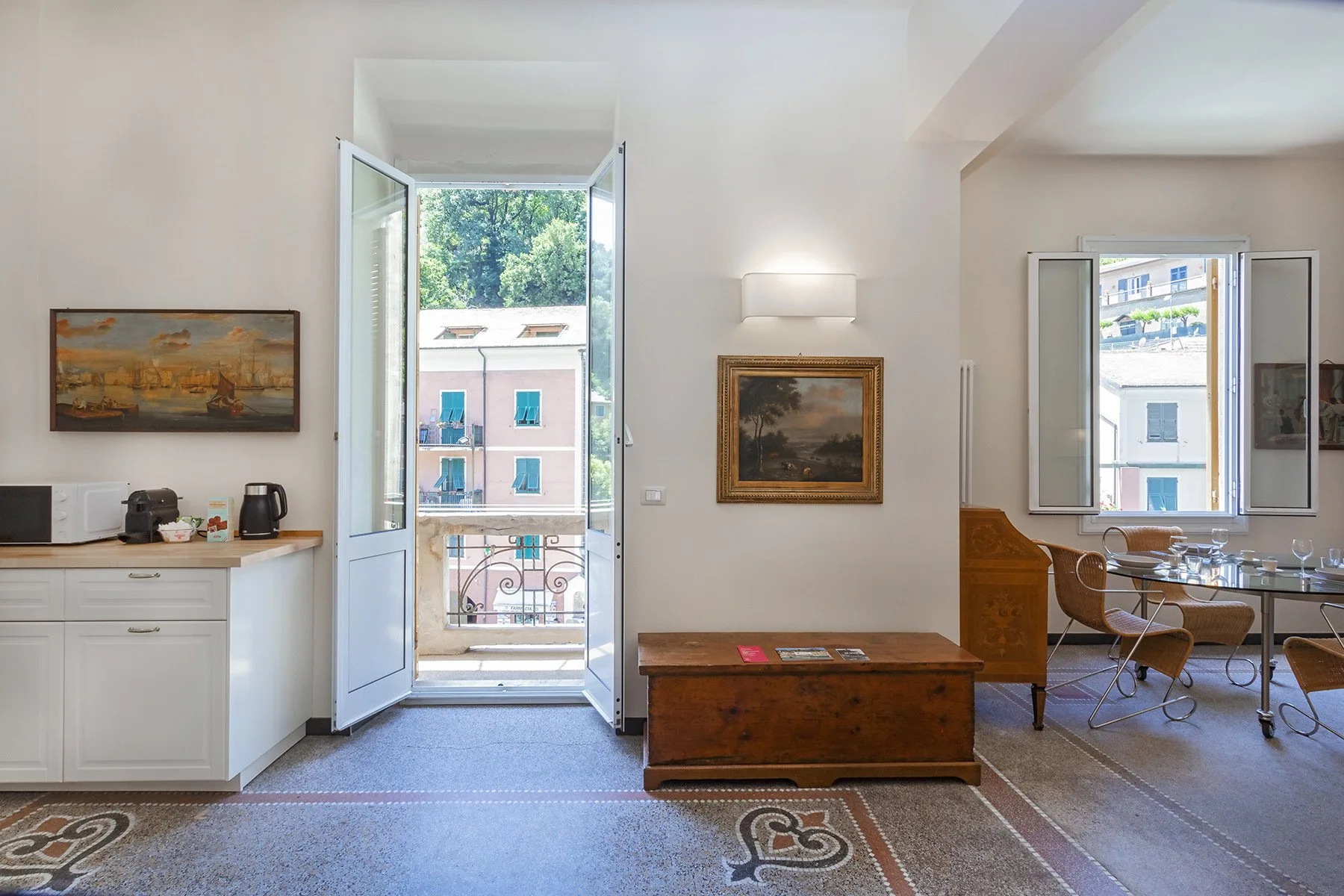 Una habitación luminosa con puertas abiertas al balcón, un baúl de madera y cuadros.