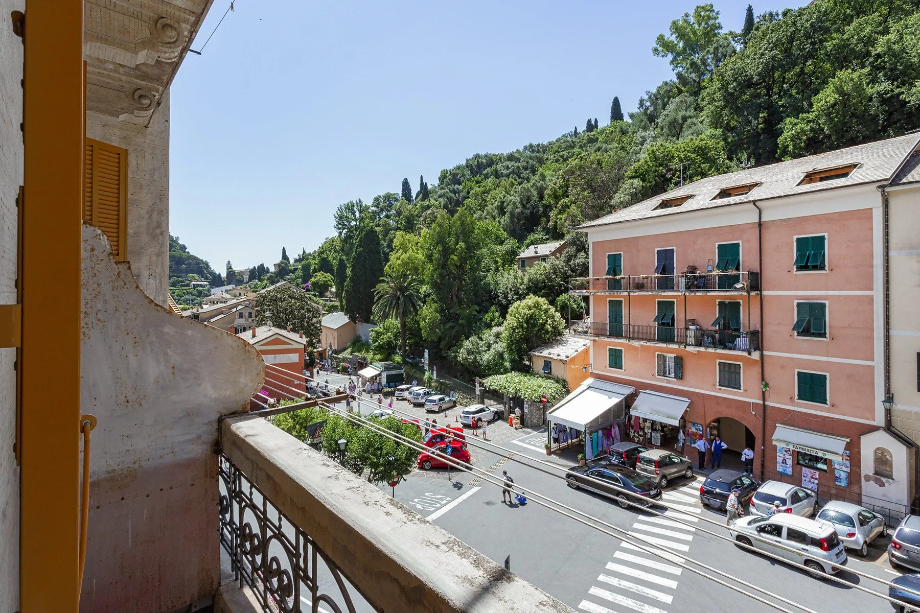 A balcony overlooks a street with a pink building and a green hillside.