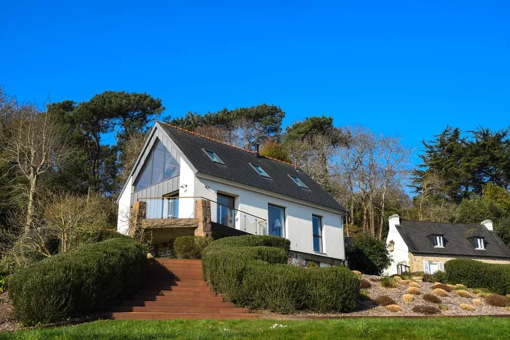 A white house with a dark pitched roof sits among trees and landscaped shrubs.