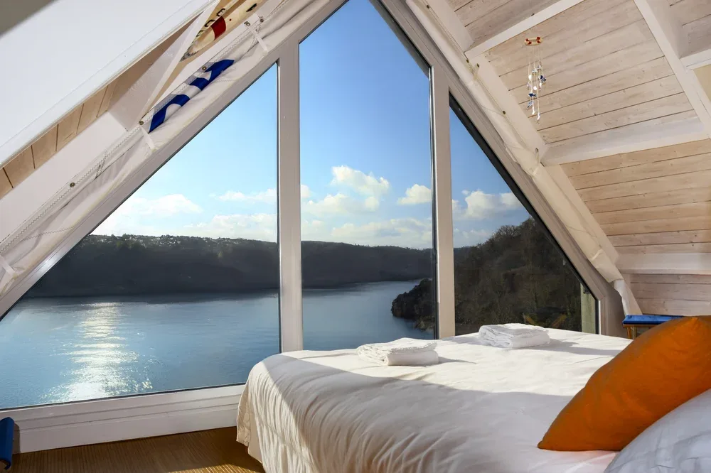 A bedroom with a bed beneath a large triangular window overlooking a lake.