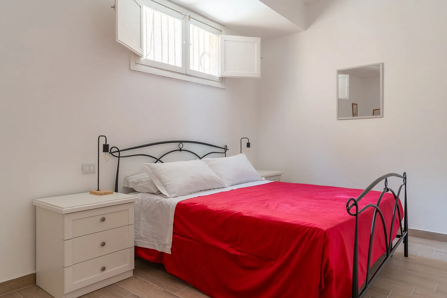 Un dormitorio con una cama de metal negro, ropa de cama blanca y una colcha roja brillante.