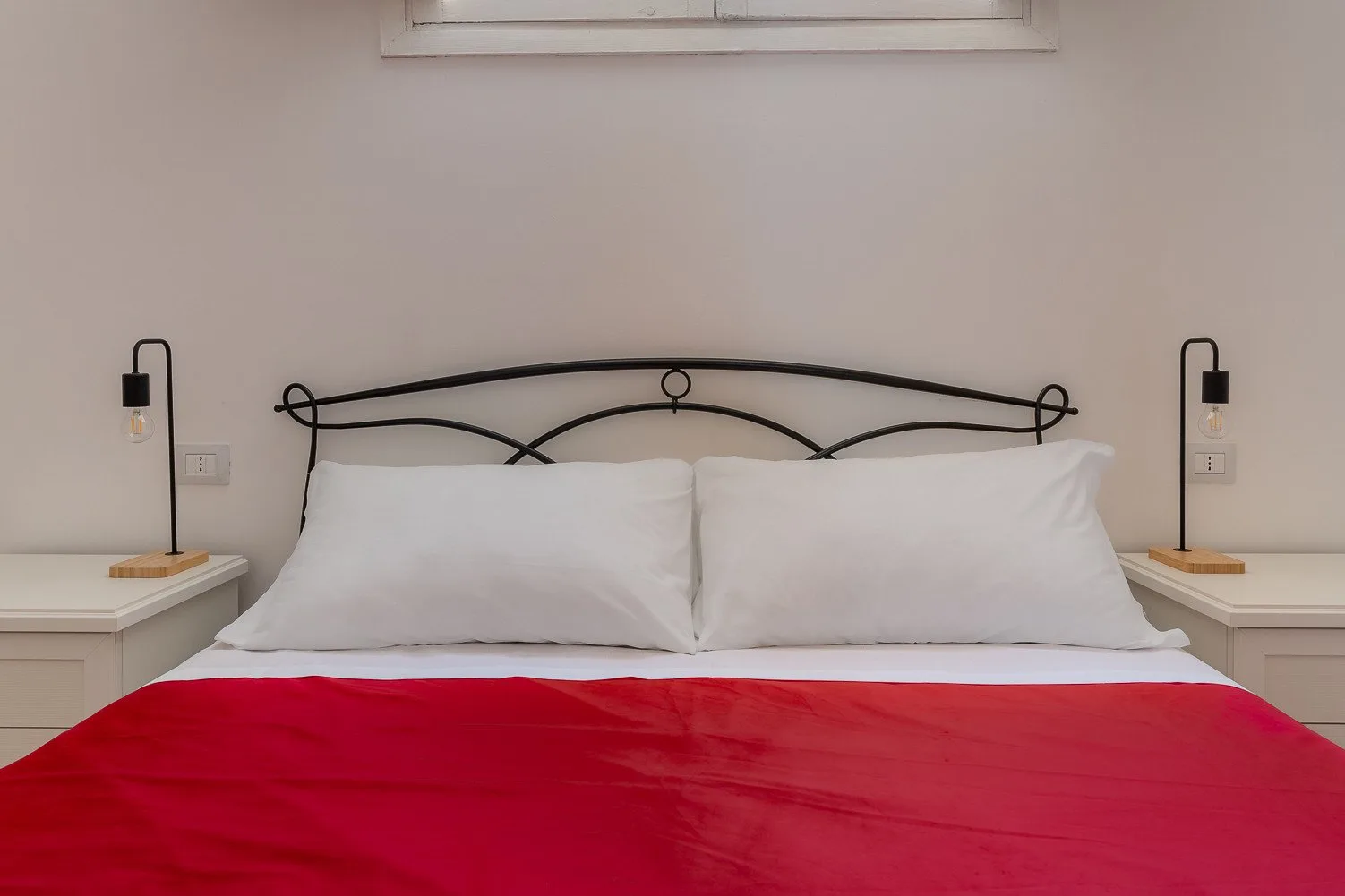 Un dormitorio con una cama de estructura metálica, almohadas blancas y una colcha roja.
