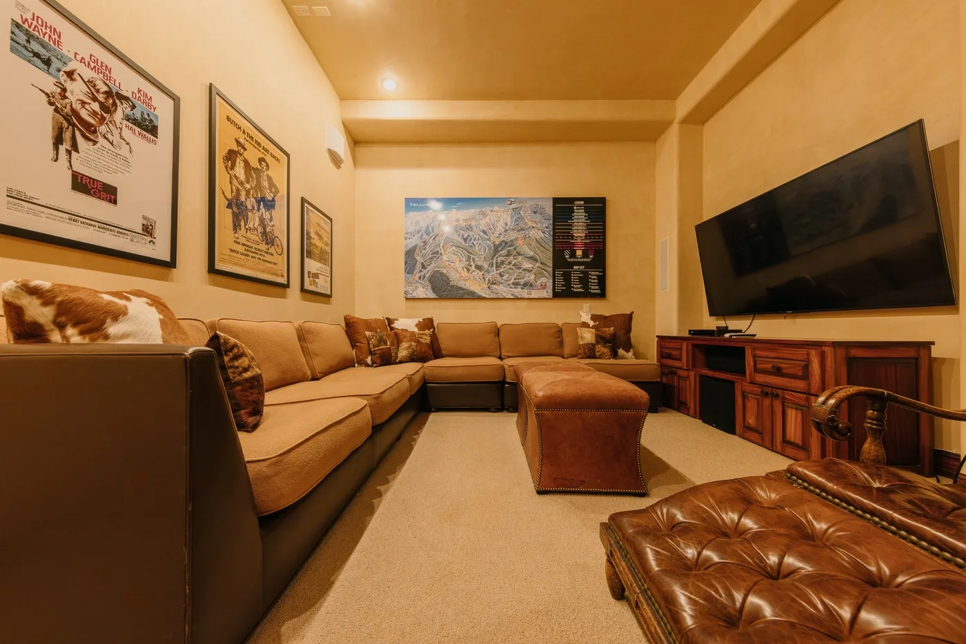 A warm-toned living room with sectional seating, an ottoman, and a wall-mounted television.