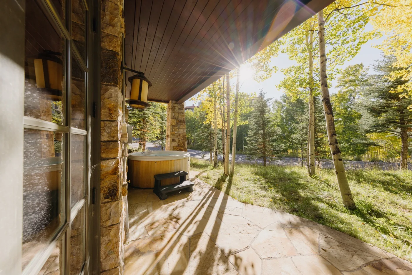 A covered stone patio with a hot tub overlooks a wooded landscape.