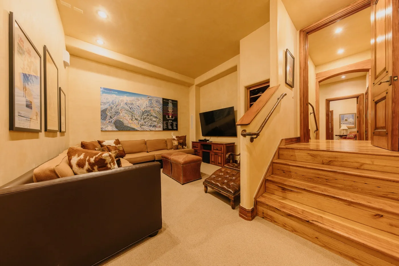 A warm-toned living room with seating, framed artwork, and wooden stairs.